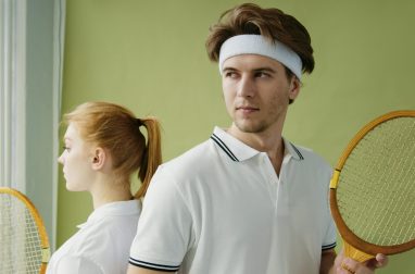 Young man and woman in sportswear holding vintage squash rackets against a green background.